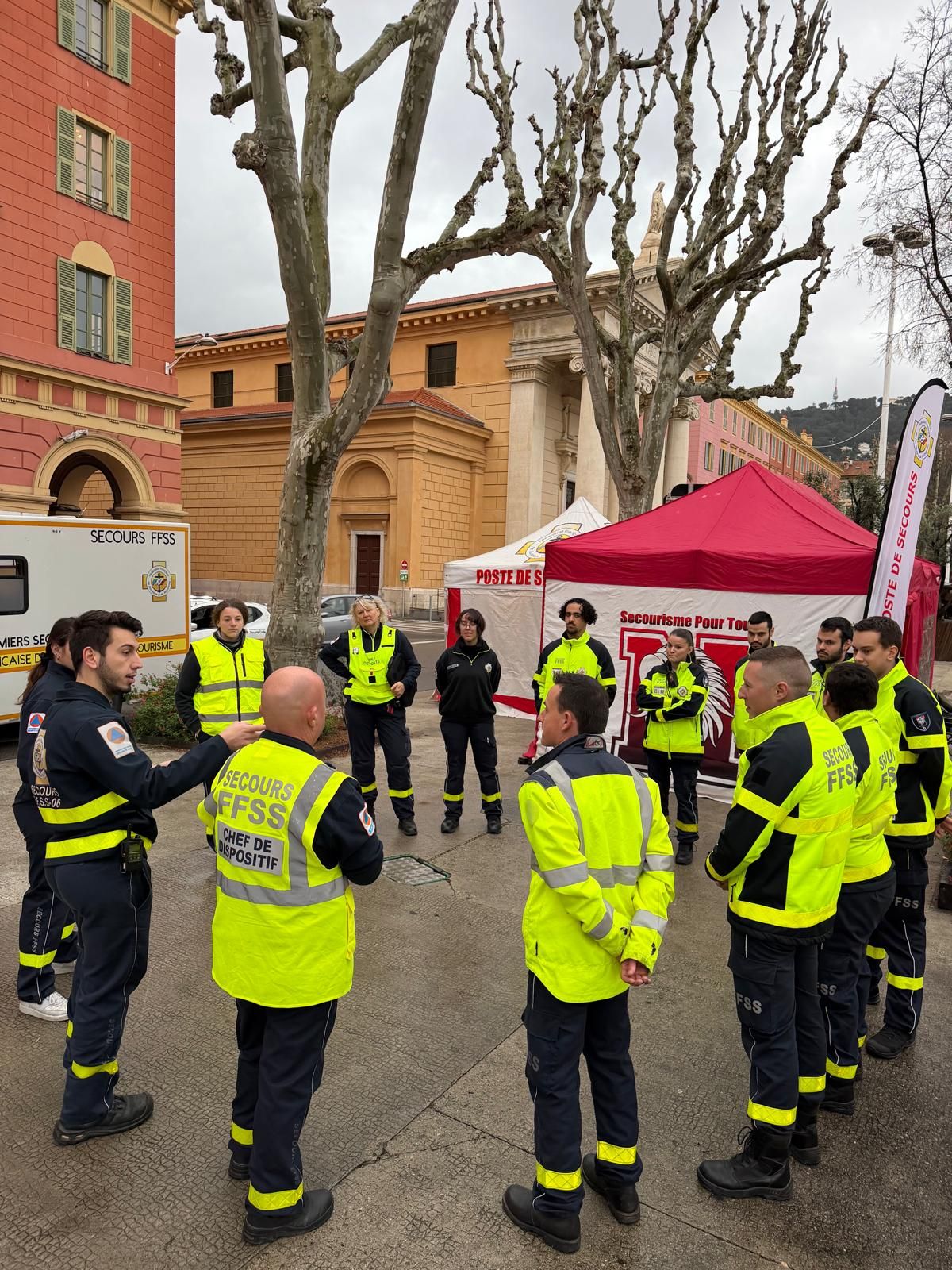 Une quinzaine de secouristes en cercle autour du chef de dispositif pour le briefing, devant la tente du poste de secours et le véhicule de premiers secours.