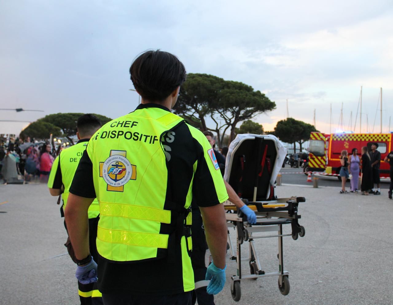 Un chef de dispositif de dos, gants aux mains, poussant un brancard vers l'ambulance des sapeurs-pompiers stationnée près du public.