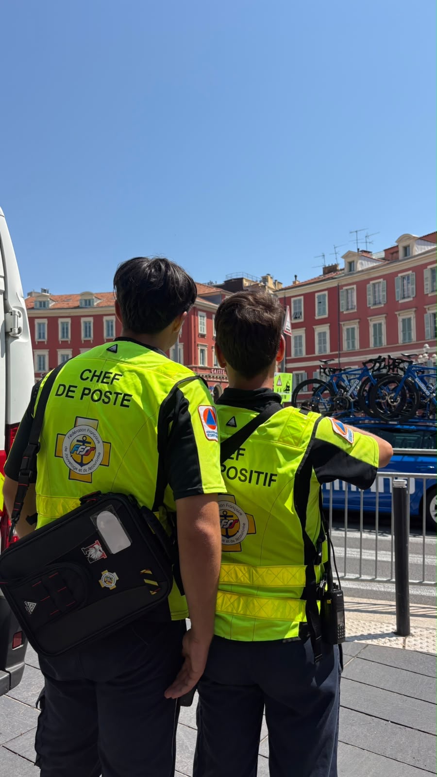 Un chef de poste et un chef de dispositif, de dos en chasuble jaune FFSS, observant le départ d'une épreuve cycliste place Masséna à Nice.