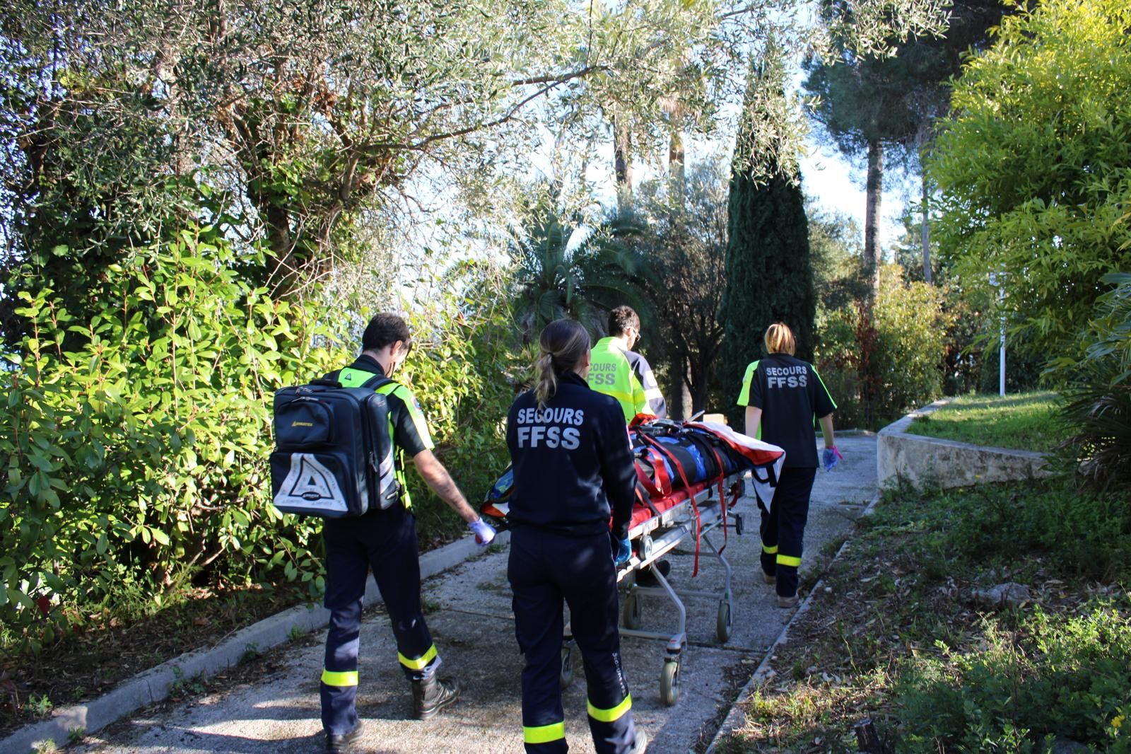Trois secouristes évacuant une victime sur un brancard le long d'une allée boisée, matériel d'intervention porté à la main.