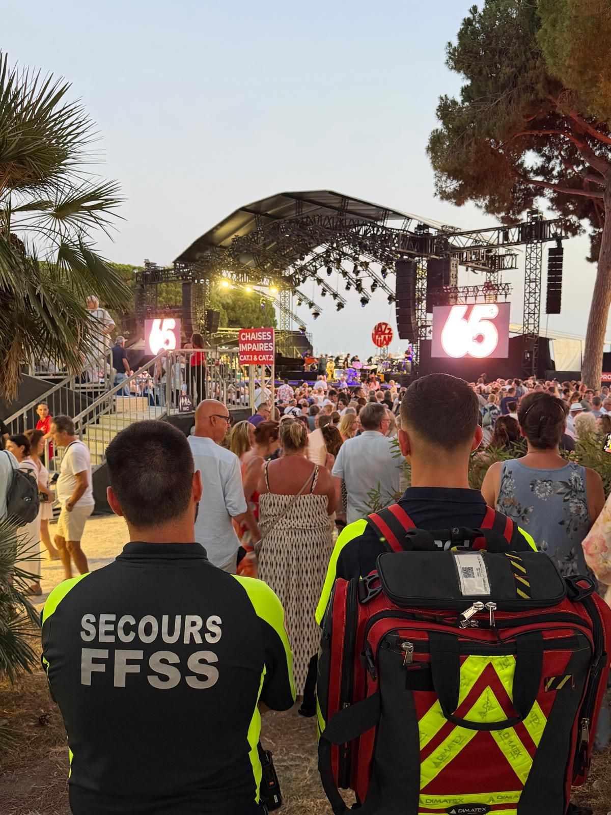 Deux secouristes sac au dos au milieu du public, face à la scène éclairée d'un concert en plein air à Juan-les-Pins.