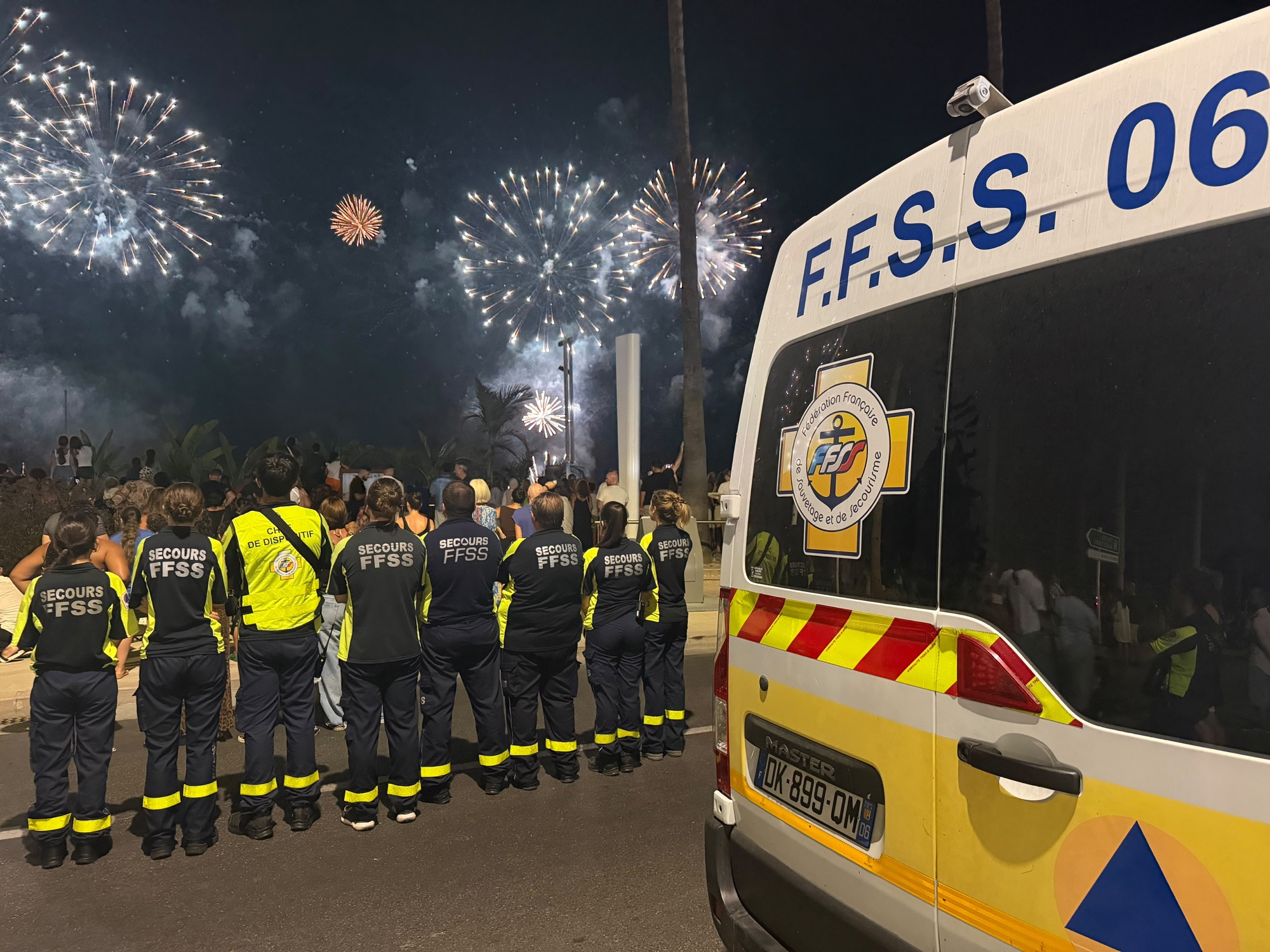 Une dizaine de secouristes en veste « Secours FFSS », de dos face au feu d'artifice, alignés le long du véhicule de premiers secours FFSS 06.