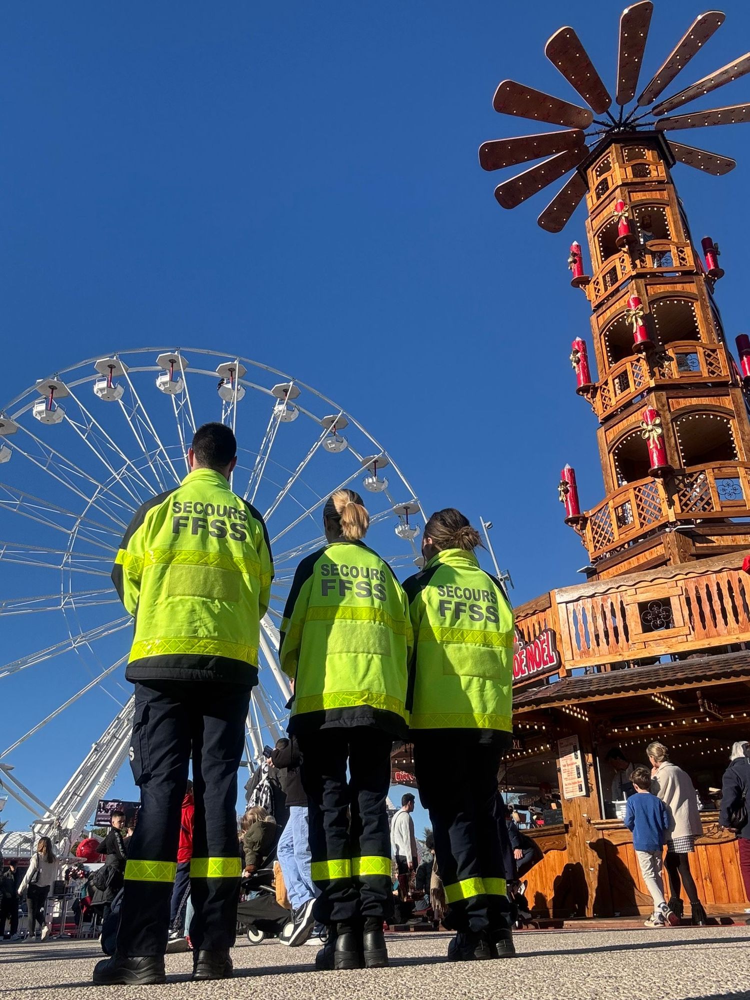 Trois secouristes de dos devant la grande roue et la pyramide de bois illuminée du marché de Noël, sous un ciel bleu.