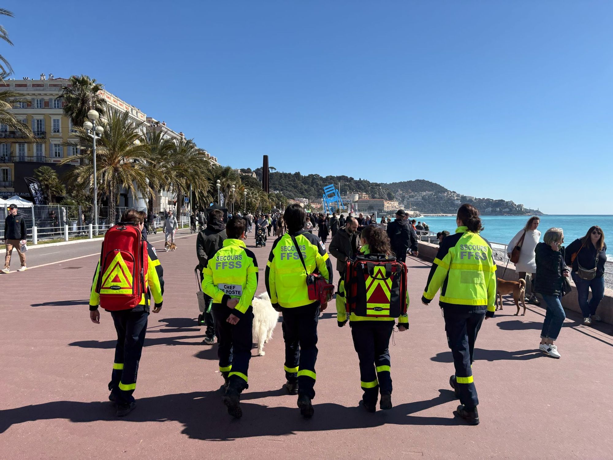 Cinq secouristes en veste haute visibilité remontant la Promenade des Anglais au milieu des passants, la mer et les palmiers en arrière-plan.
