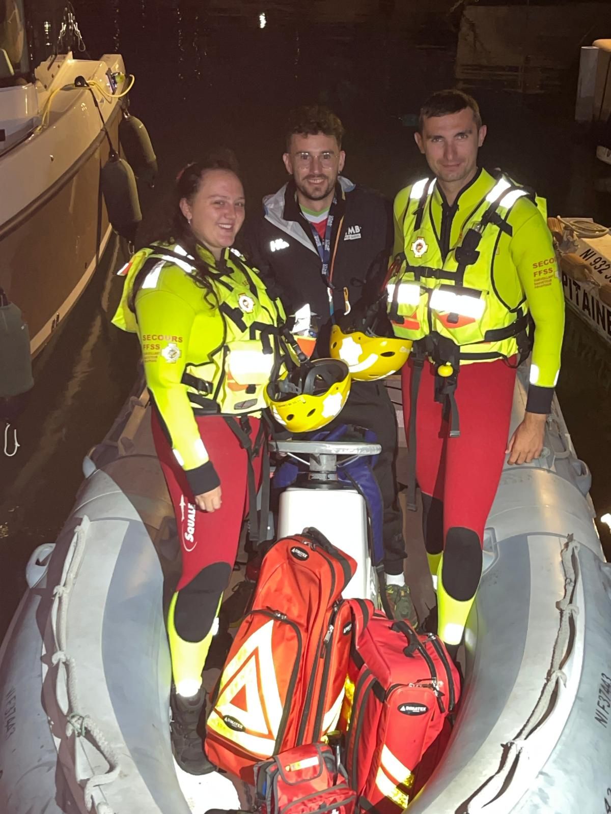Trois sauveteurs en combinaison de sauvetage debout dans un bateau semi-rigide amarré de nuit, sacs de secours et casques à bord.