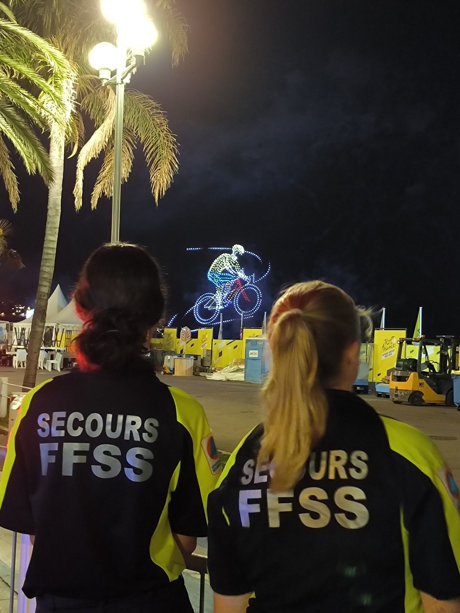 Deux secouristes de dos sur la Promenade des Anglais, face au spectacle de drones lumineux dessinant un cycliste dans le ciel de Nice.