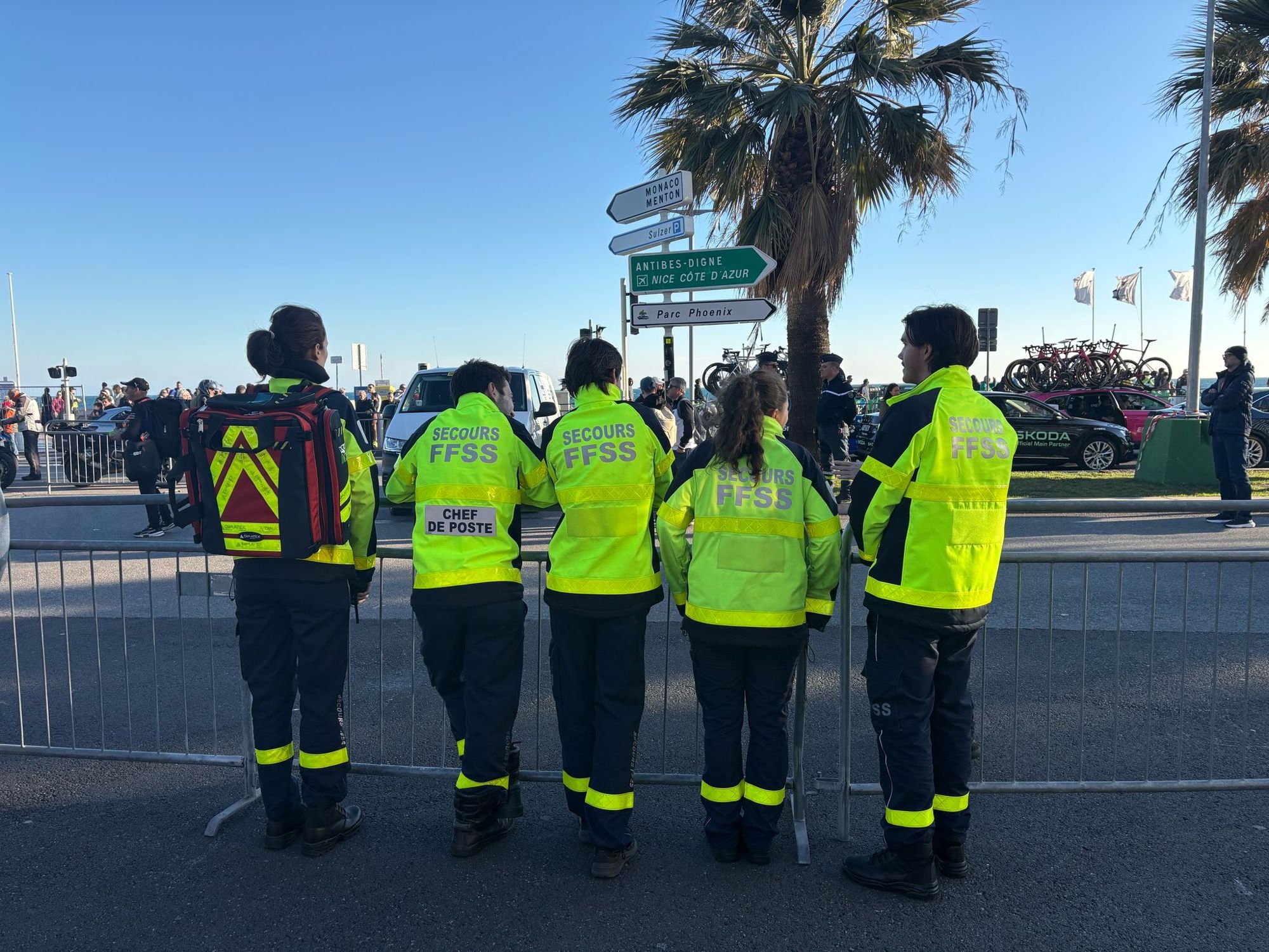 Cinq secouristes en veste haute visibilité alignés derrière les barrières, dos au photographe, au départ d'une étape du Tour de France à Nice.