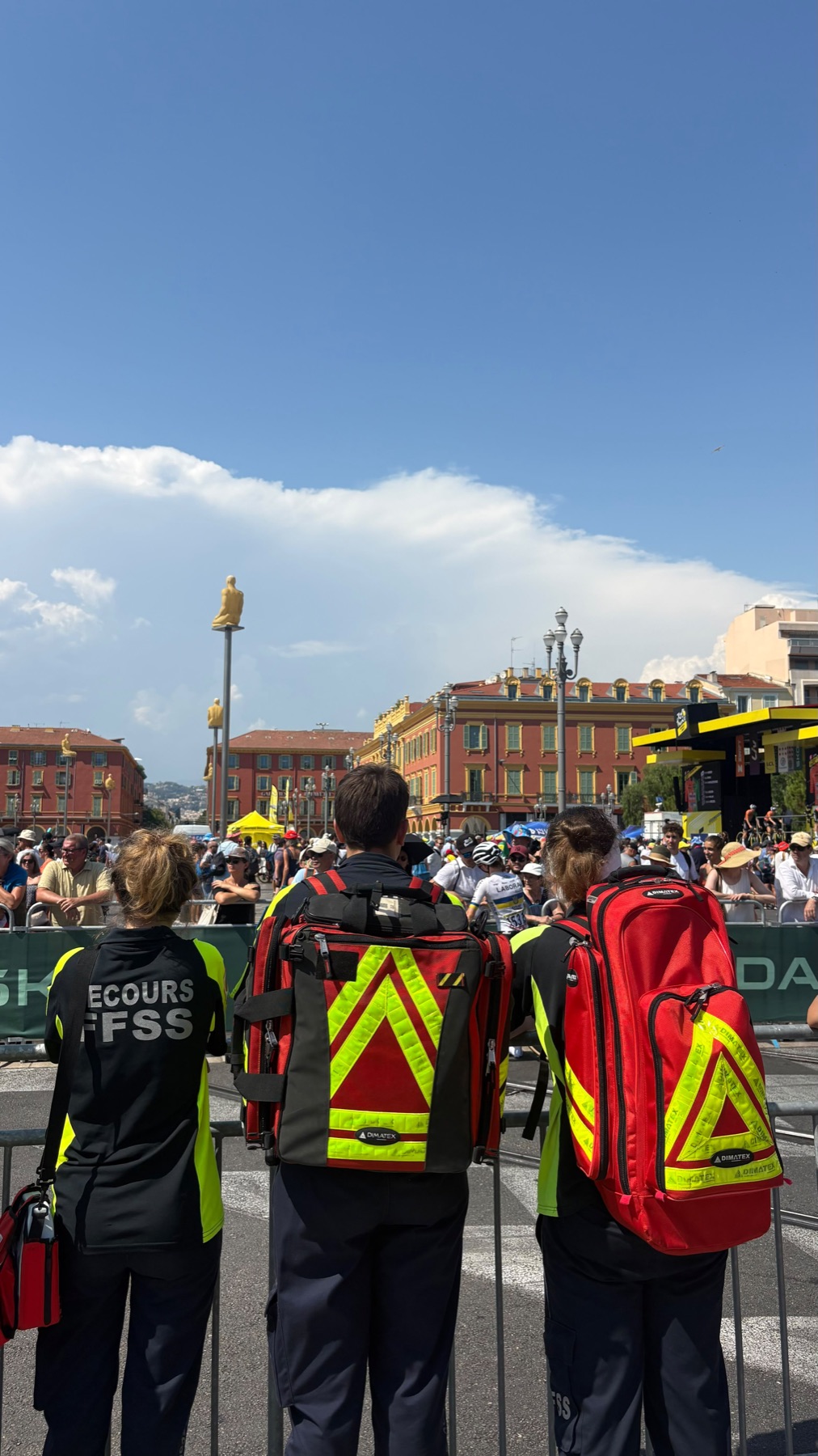 Trois secouristes de Secourisme Pour Tous en tenue FFSS, de dos derrière les barrières, face au passage des coureuses place Masséna à Nice.