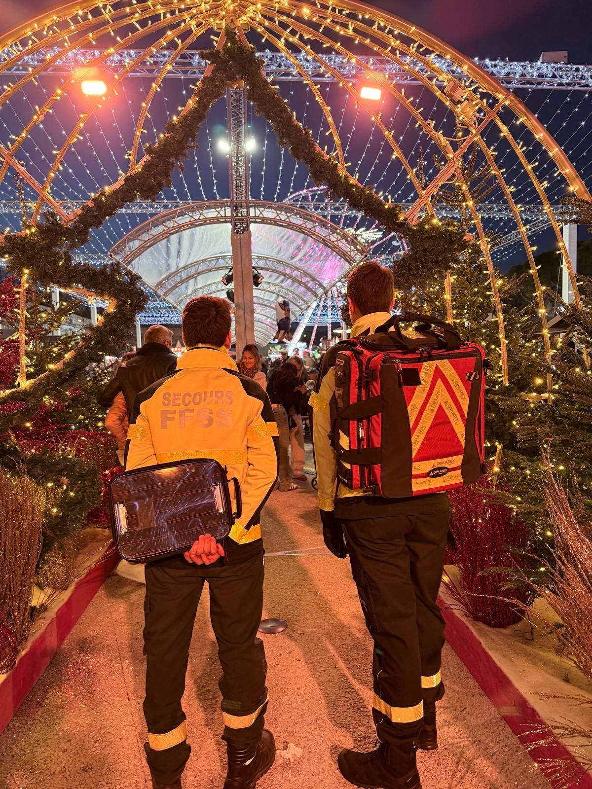 Deux secouristes marchant sous les arches lumineuses d'un village de Noël, sac d'intervention et lot de brancardage à la main.